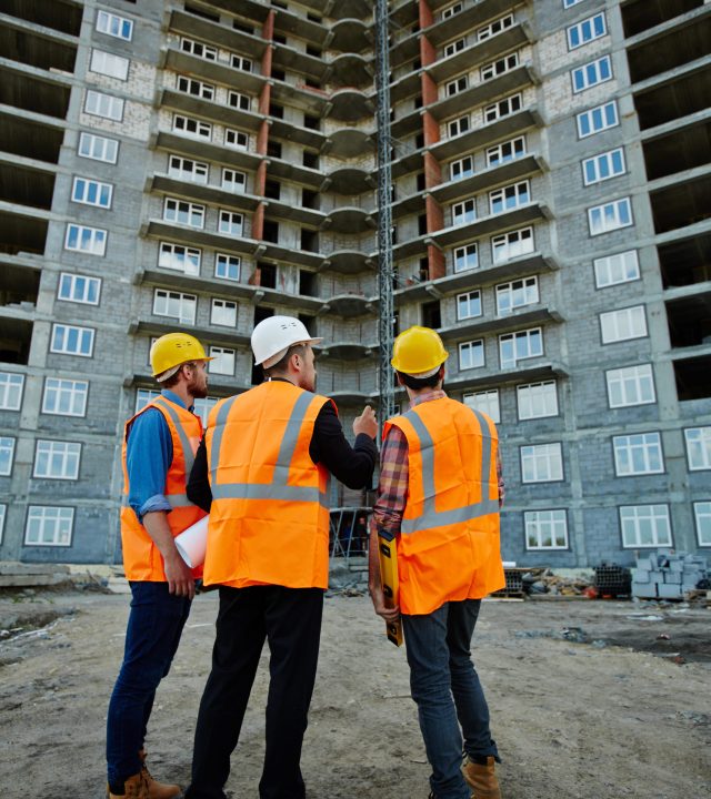 Back view portrait of three workmen wearing reflective orange vests and hard hats standing on construction site against unfinished apartment building, discussing it